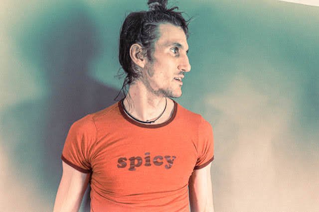 Side profile of a young man with dark, messy hair, wearing a red t-shirt with the word 'spicy' on it, against a green background.