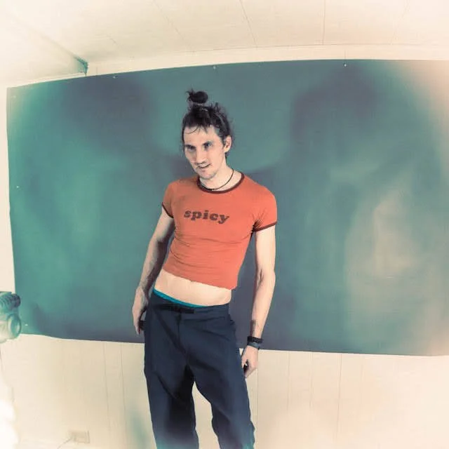 A person with a top knot hairstyle wearing a red crop top that says 'spicy', black pants, and a black choker, standing in front of a teal background.