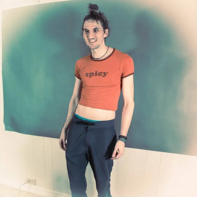 Person with curly hair in a high bun, wearing a red cropped t-shirt with 'spicy' written on it, dark pants, standing in front of a green backdrop, smiling at the camera.