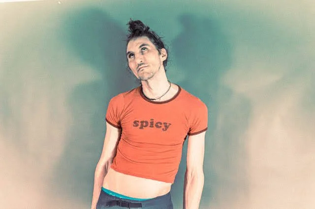 Person with dark hair tied up, wearing a red shirt with the word 'spicy' on it, standing against a greenish background with a contemplative expression.
