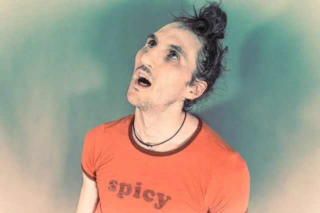 A man with long hair tied up, wearing an orange shirt with the word 'spicy' on it, looking upwards with a surprised or confused expression.