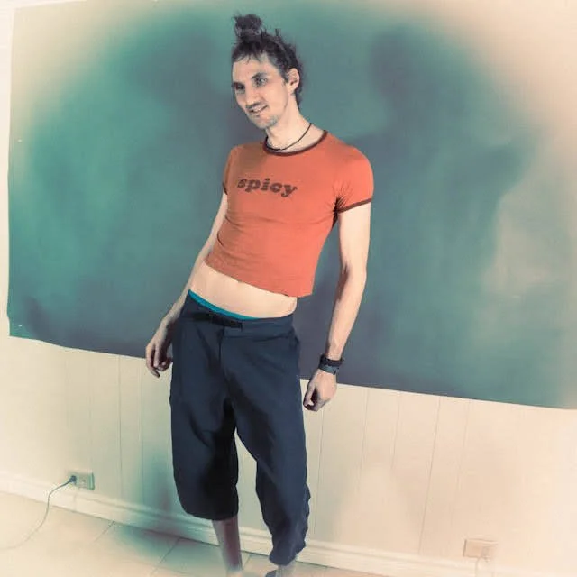 A young person with long hair tied in a bun, wearing a cropped red t-shirt with the word 'spicy' on it, black wristwatch, and dark pants, standing indoors against a turquoise background.