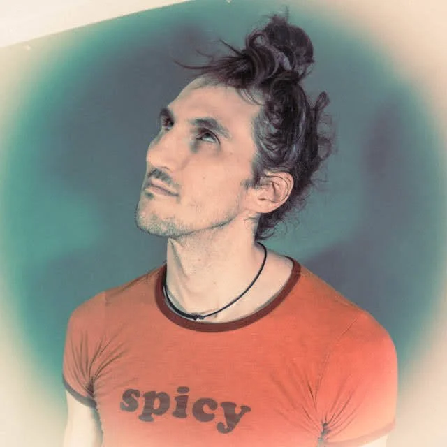 A man with long, curly hair tied up in a topknot, wearing an orange t-shirt with the word 'spicy' printed on it, looking upwards against a gradient background.