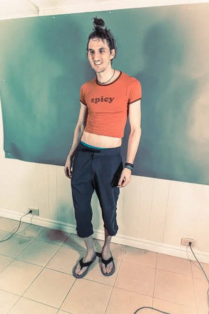 Young woman with a bun hair style wearing a red crop top with 'spicy' written on it, navy blue jogger pants, flip-flops, smiling, standing indoors against a green wall.