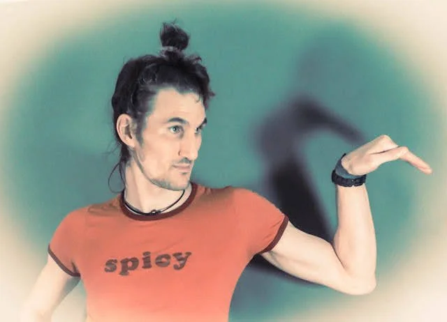 A man with black hair tied in a bun, wearing an orange t-shirt with the word "spicy," poses with his arm bent and hand facing downward against a green background.