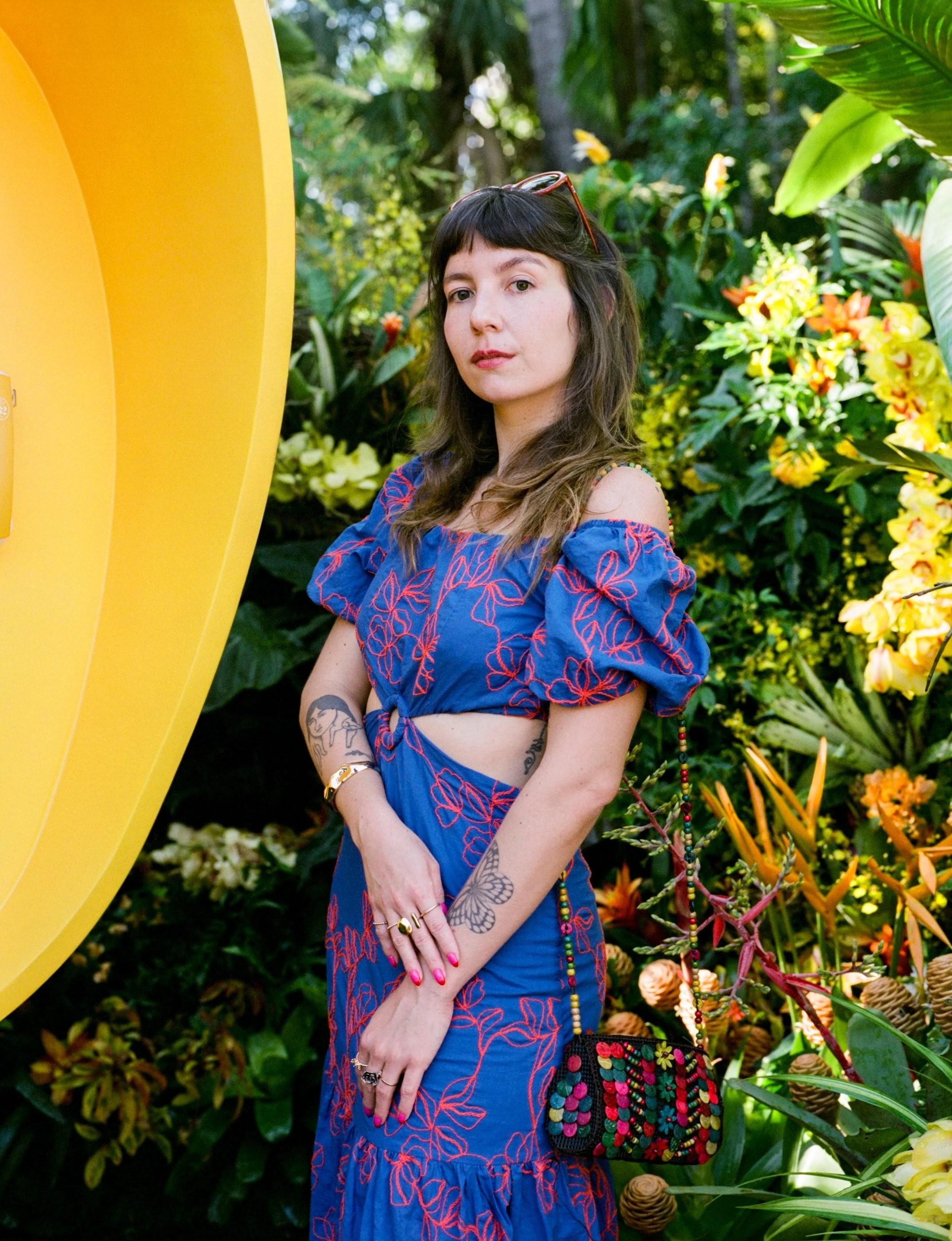 A woman with long brown hair and light skin standing in a lush garden with green foliage and yellow flowers. She is wearing a blue dress with red embroidered patterns, jewelry, and sunglasses on her head. She has tattoos on her arms and hands, and is