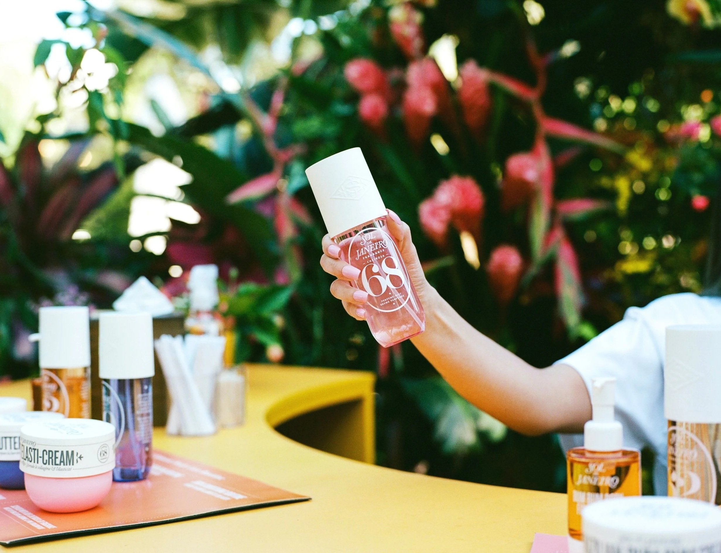 A person holding a pink spray bottle with white accents at an outdoor table with skincare products, including creams and bottles, on a yellow surface, with a background of lush green and pink tropical plants.