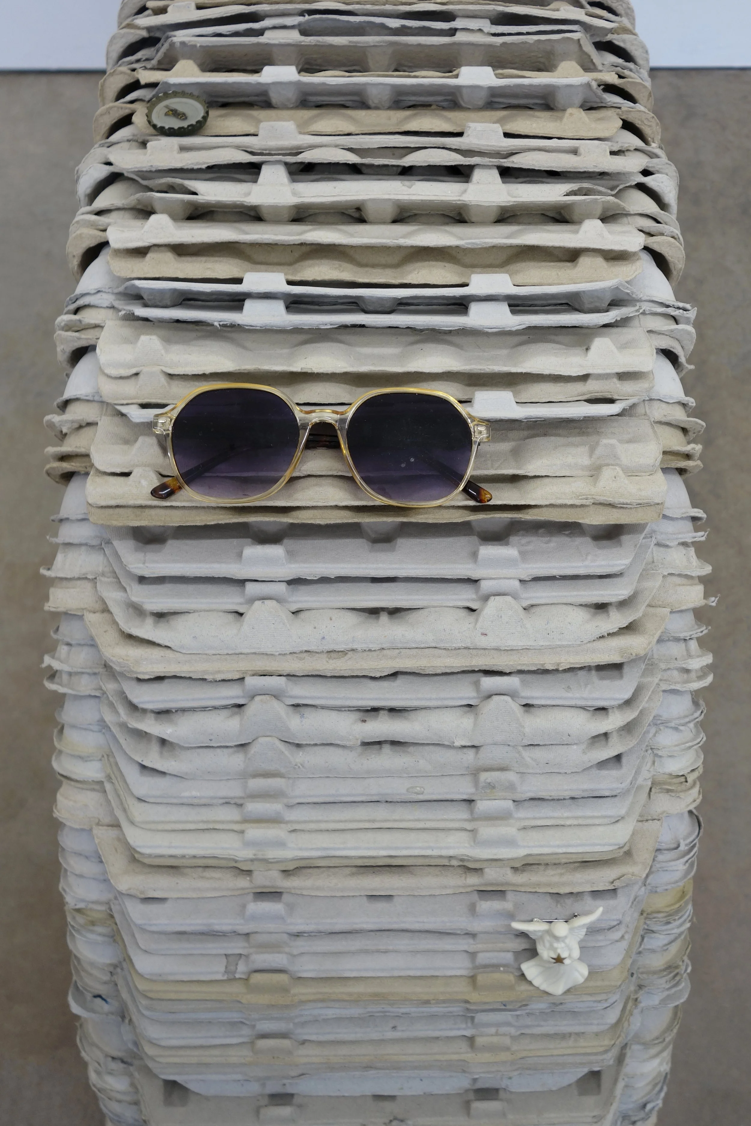   Investment , 2026. egg cartons, nail, cast and hand-painted epoxy resin, shell casing, yellow jacket and epoxy resin in bottle cap, sunglasses, angel pin, door key, and steel, 19 x 46 x 12 in (48.3 x 116.8 x 30.5 cm) 