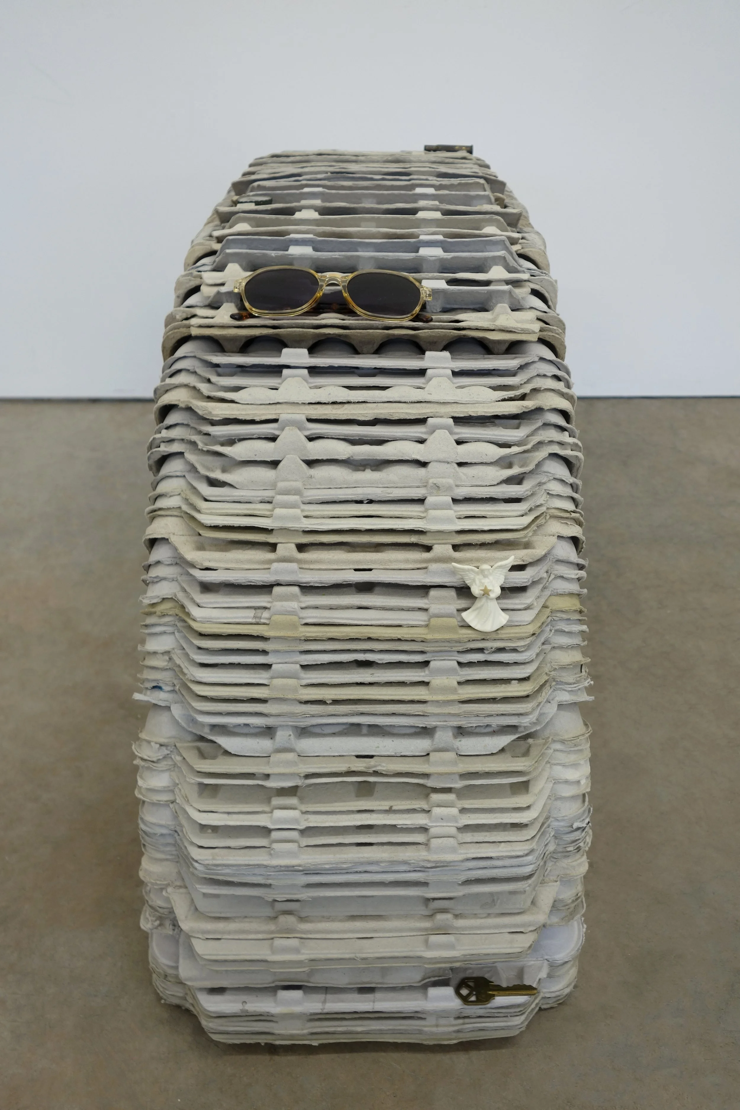   Investment , 2026. egg cartons, nail, cast and hand-painted epoxy resin, shell casing, yellow jacket and epoxy resin in bottle cap, sunglasses, angel pin, door key, and steel, 19 x 46 x 12 in (48.3 x 116.8 x 30.5 cm) 