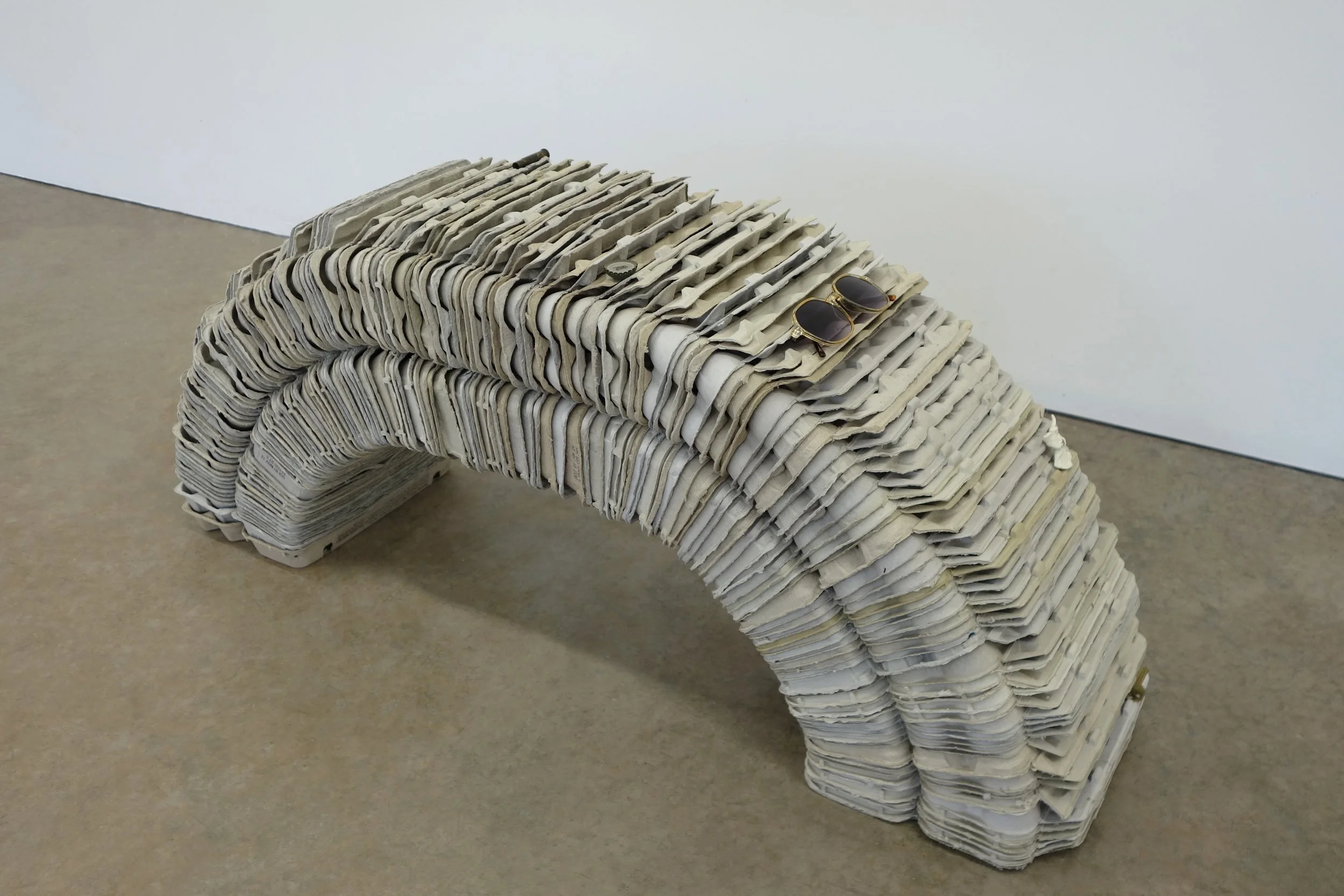   Investment , 2026. egg cartons, nail, cast and hand-painted epoxy resin, shell casing, yellow jacket and epoxy resin in bottle cap, sunglasses, angel pin, door key, and steel, 19 x 46 x 12 in (48.3 x 116.8 x 30.5 cm) 