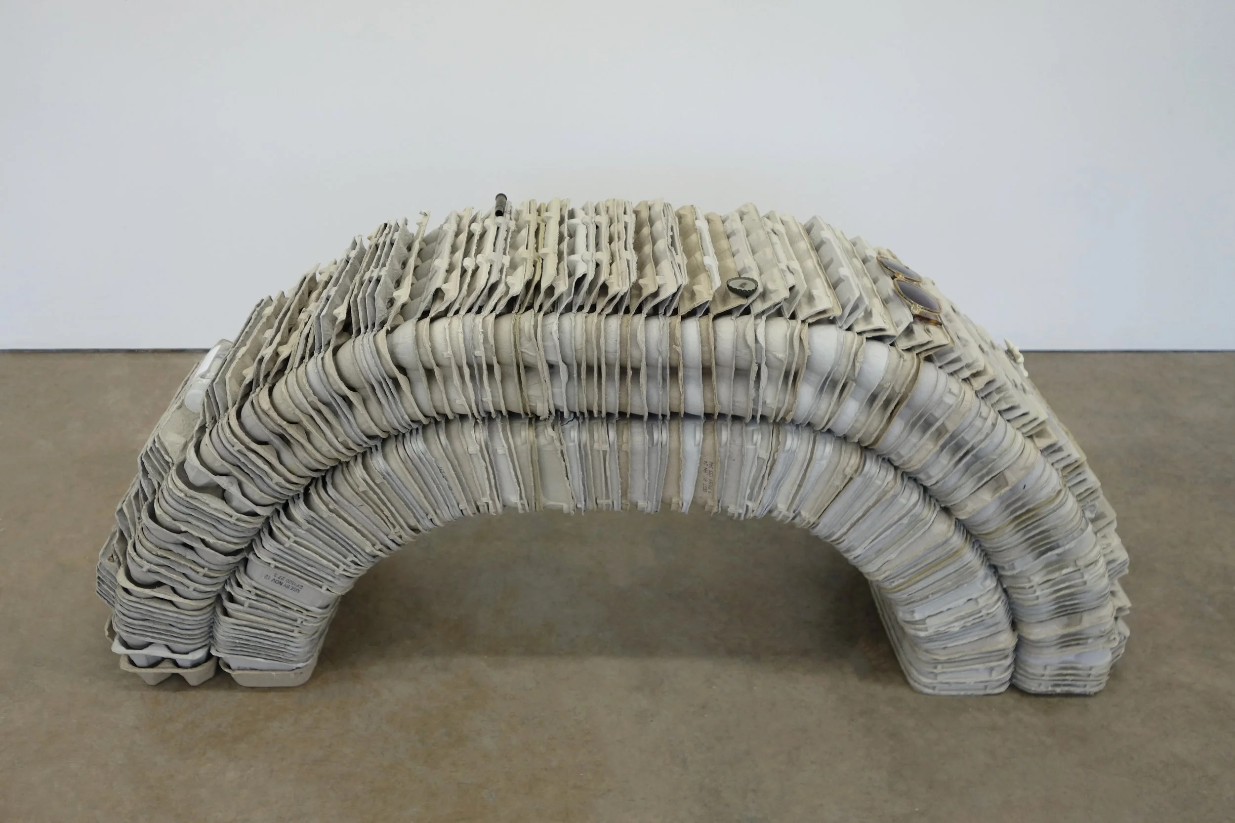   Investment , 2026. egg cartons, nail, cast and hand-painted epoxy resin, shell casing, yellow jacket and epoxy resin in bottle cap, sunglasses, angel pin, door key, and steel, 19 x 46 x 12 in (48.3 x 116.8 x 30.5 cm) 