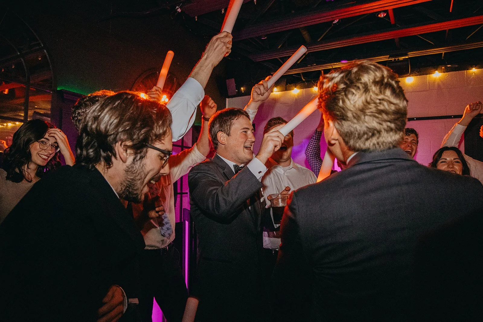 Groom wedding party and guests dance with vibrantly colored glowsticks against the brilliant lights of the Revel Center in Grand Rapids, Michigan