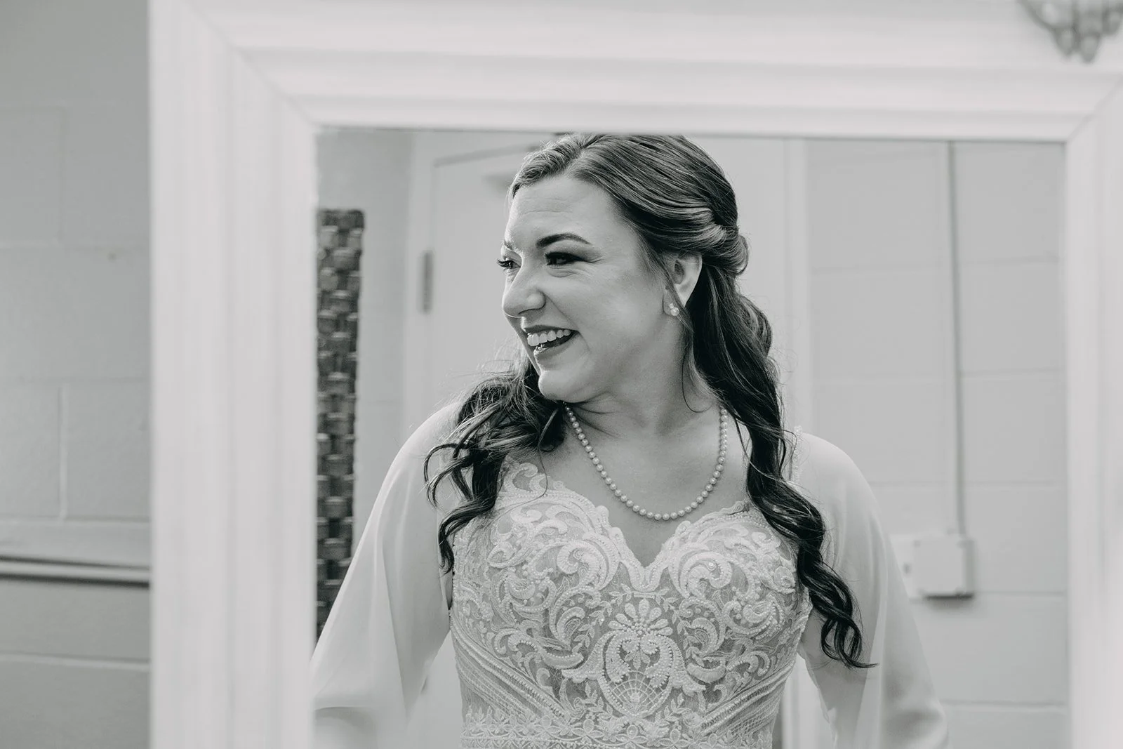 A bride smiles as she sees herself fully dressed for the first time in the elegant bridal suite of the Revel Center. Photo captured in black and white.