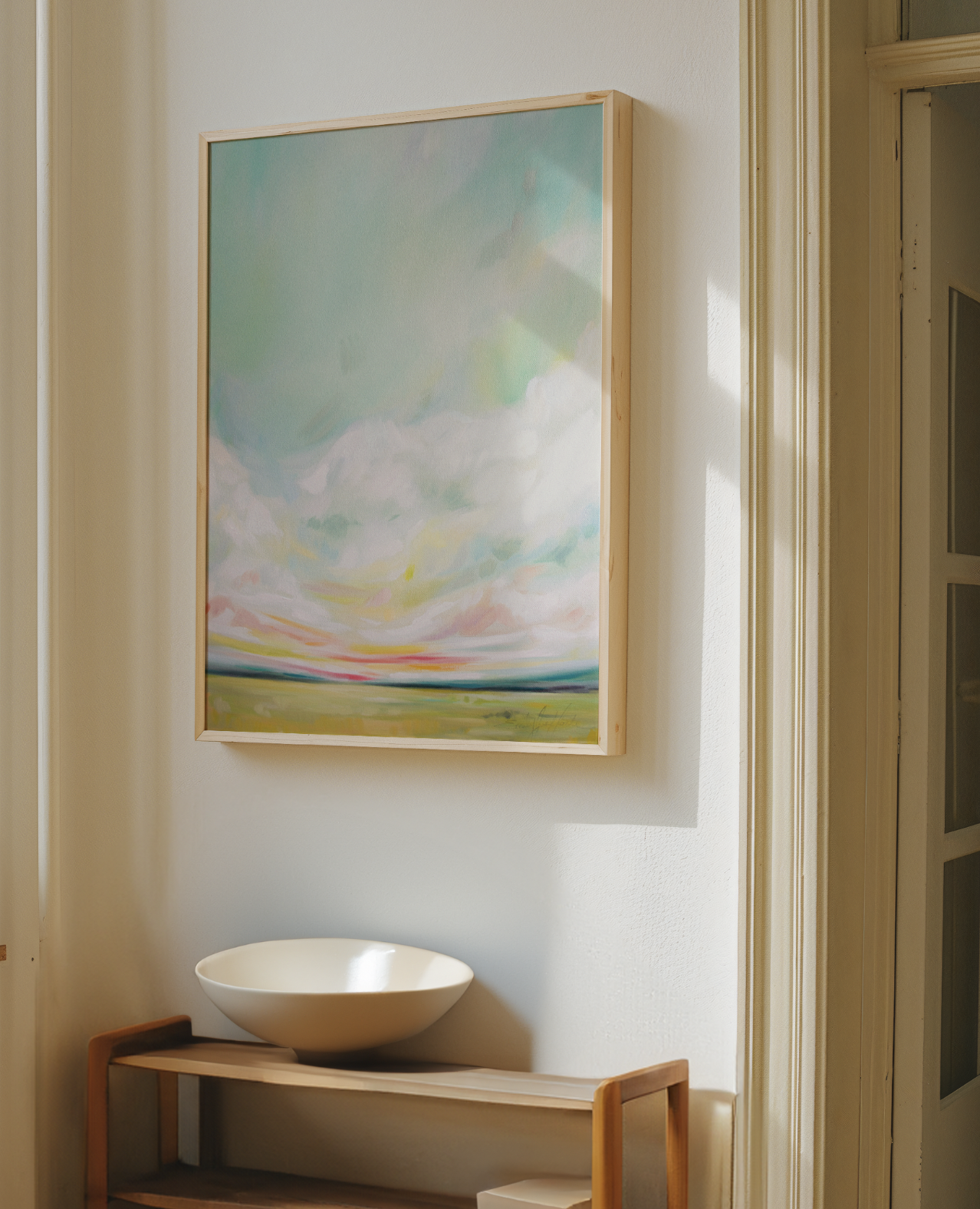 A framed landscape painting hanging on a white wall with a wooden border, above a wooden console table with a white ceramic bowl, next to a curved doorframe and window.