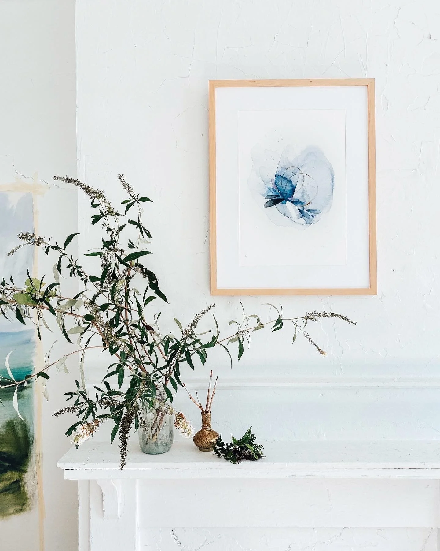 White wall with a framed watercolor painting and a white shelf holding a vase with greenery and a small decorative bottle.