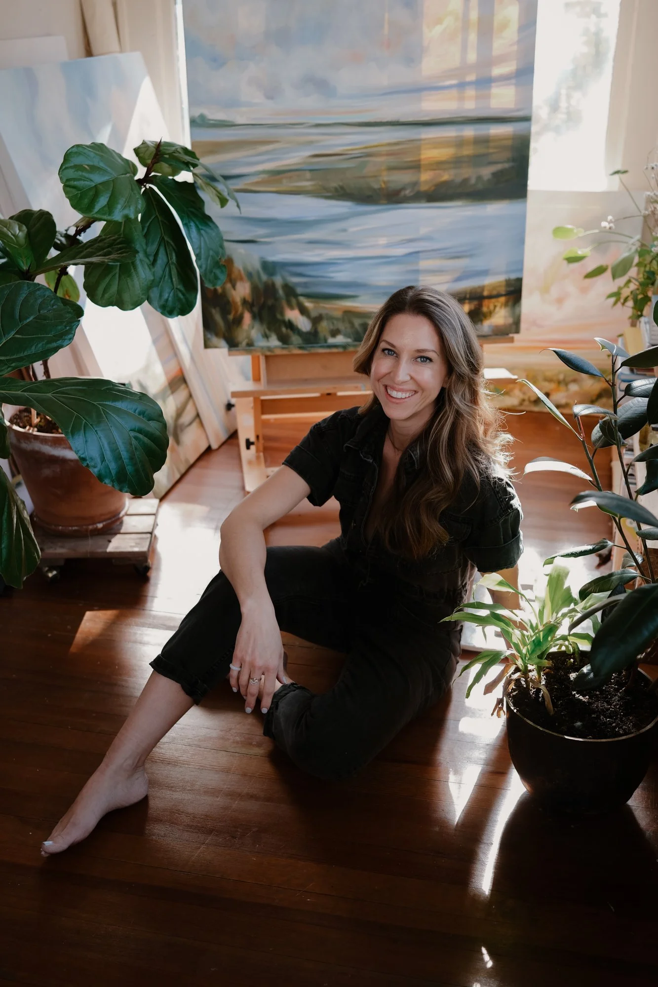 Emily sits on the floor in front of her large painting of a landscape. She's neighbored by a fiddle leaf potted plant and a few other house plants in front of the art on the easel, and she is sitting with one arm resting on her knee, smiling.