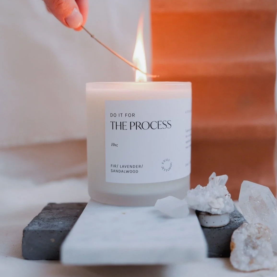 A white scented candle in a jar labeled 'Do It For The Process' with lavender and sandalwood scent, being lit with a matchstick, placed on a black and white platform surrounded by crystals.