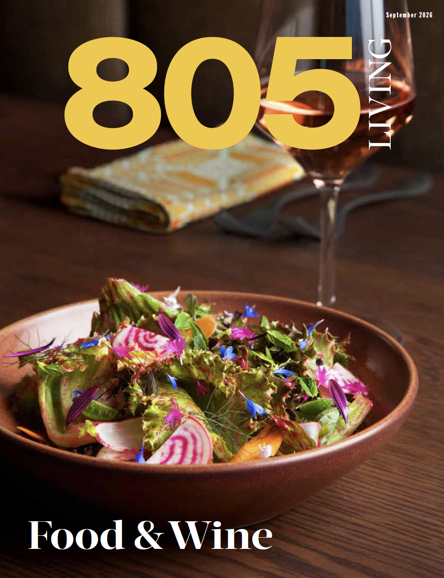 A colorful salad with edible flowers in a brown bowl, a glass of red wine, a folded patterned napkin, and a dark wooden table.