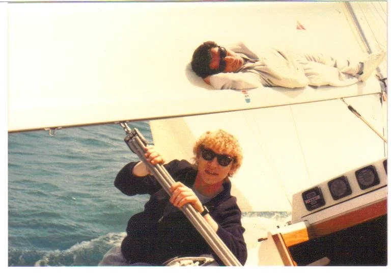 Retrato de Juan y Ramon navegando a bordo de un velero. Se les ve relajados, Juan mirando a cámara y Ramón estirado en el hueco que deja la vela mayor