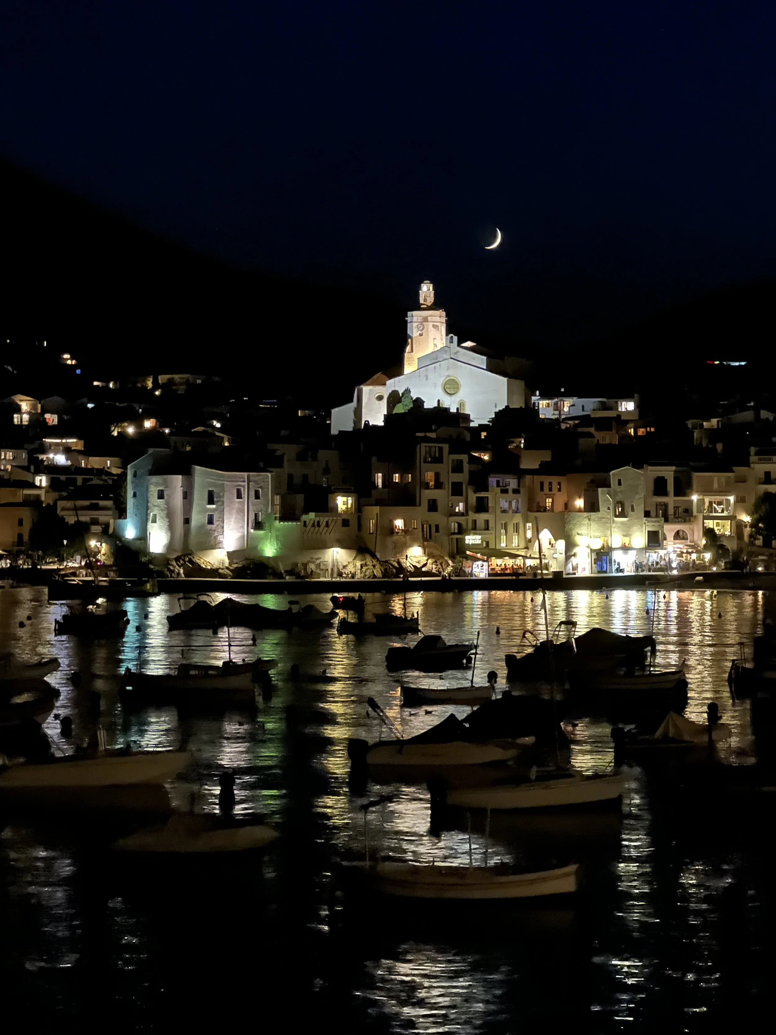 Wallpaper para celular de una vista nocturna del pueblo costero de Cadaqués iluminado bajo la luna, descargable gratuito de Viajes Magazine.
