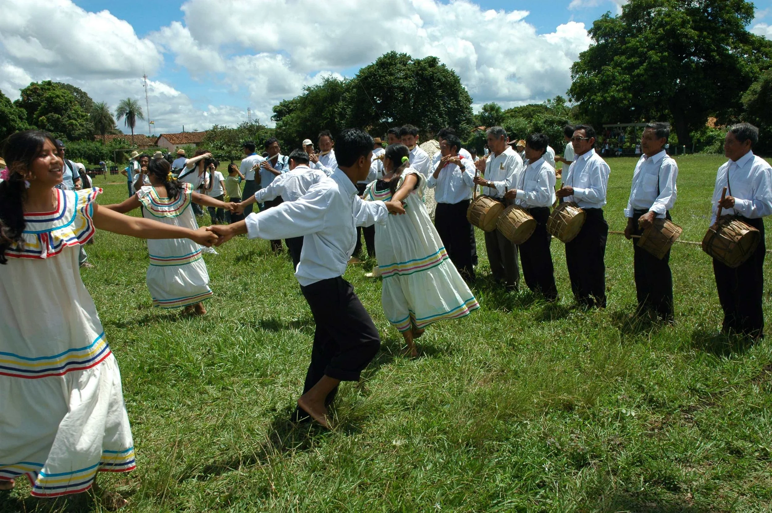 Hombres y mujeres con trajes típicos blancos bailando en círculo en un campo verde, acompañados por músicos con tambores en la Chiquitania.
