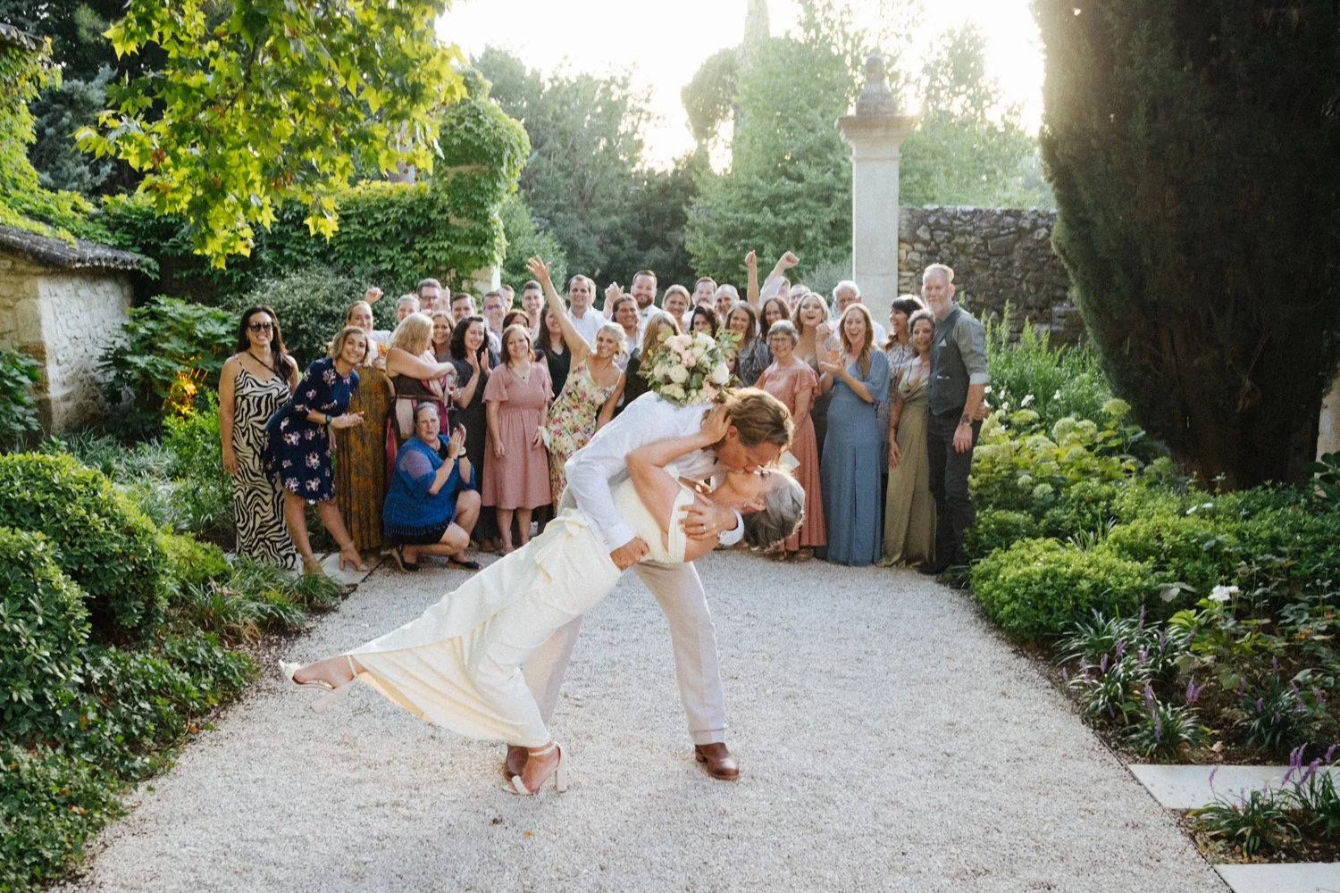A bride and groom sharing a kiss during their wedding, with the bride held horizontally, surrounded by a group of wedding guests in a garden setting at sunset.