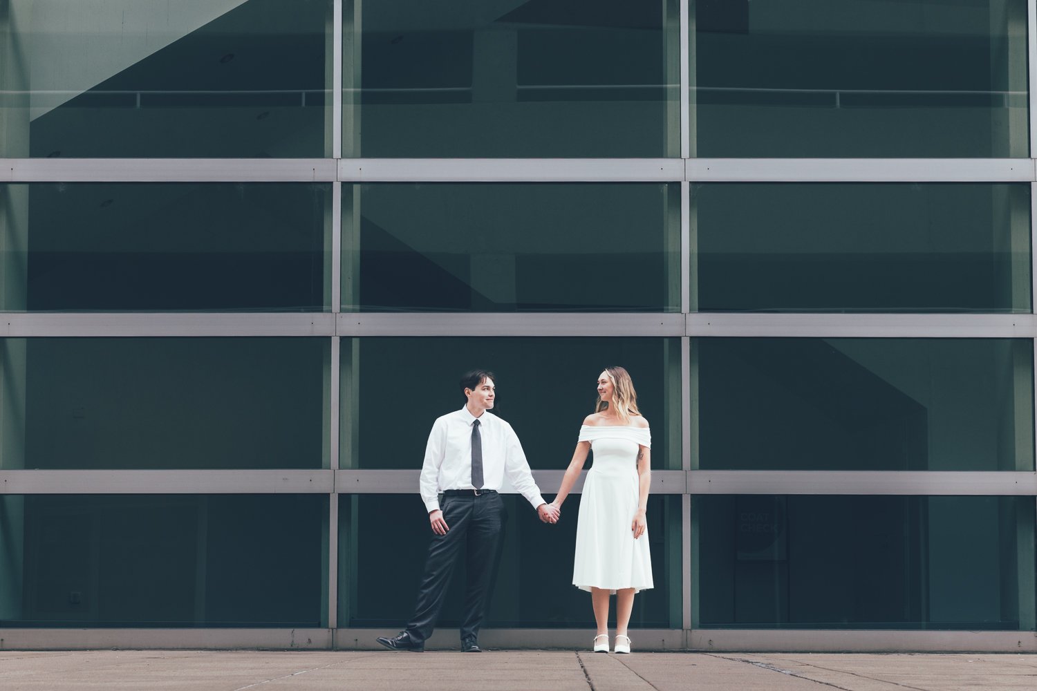 A man and woman holding hands, standing in front of a modern glass building, dressed as a groom and bride in white wedding attire.