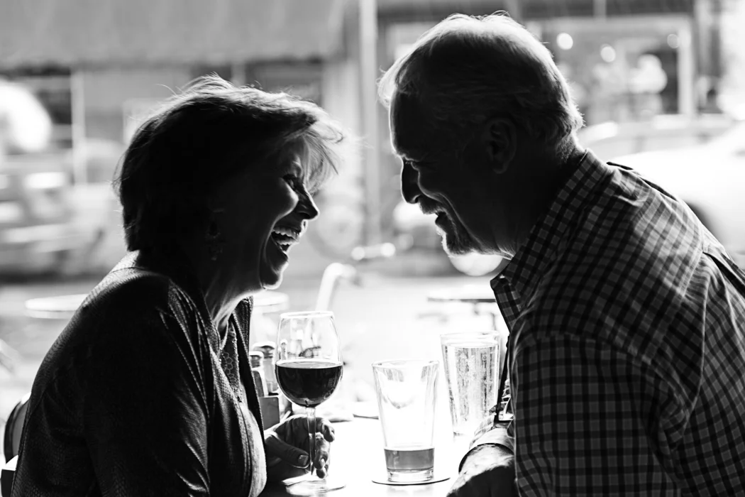 A black and white photo of a smiling woman and man with glasses of wine, sitting at a table in a restaurant or cafe, engaging in a conversation with a blurred outdoor background.