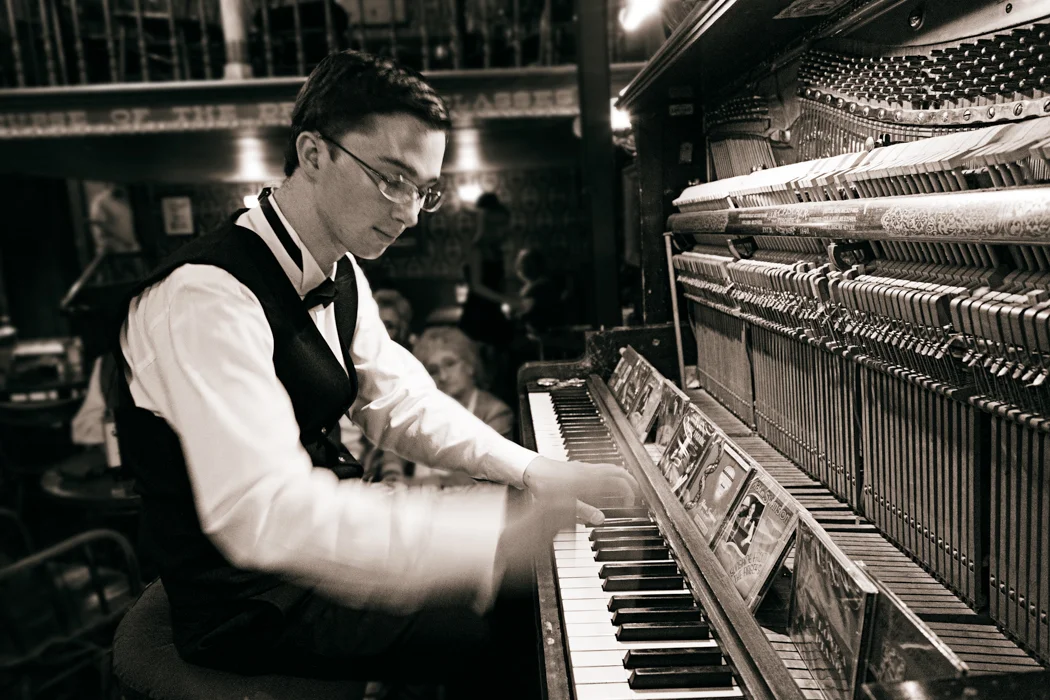 A man in a vest and bow tie playing a piano in a dimly lit, lively restaurant or bar setting.
