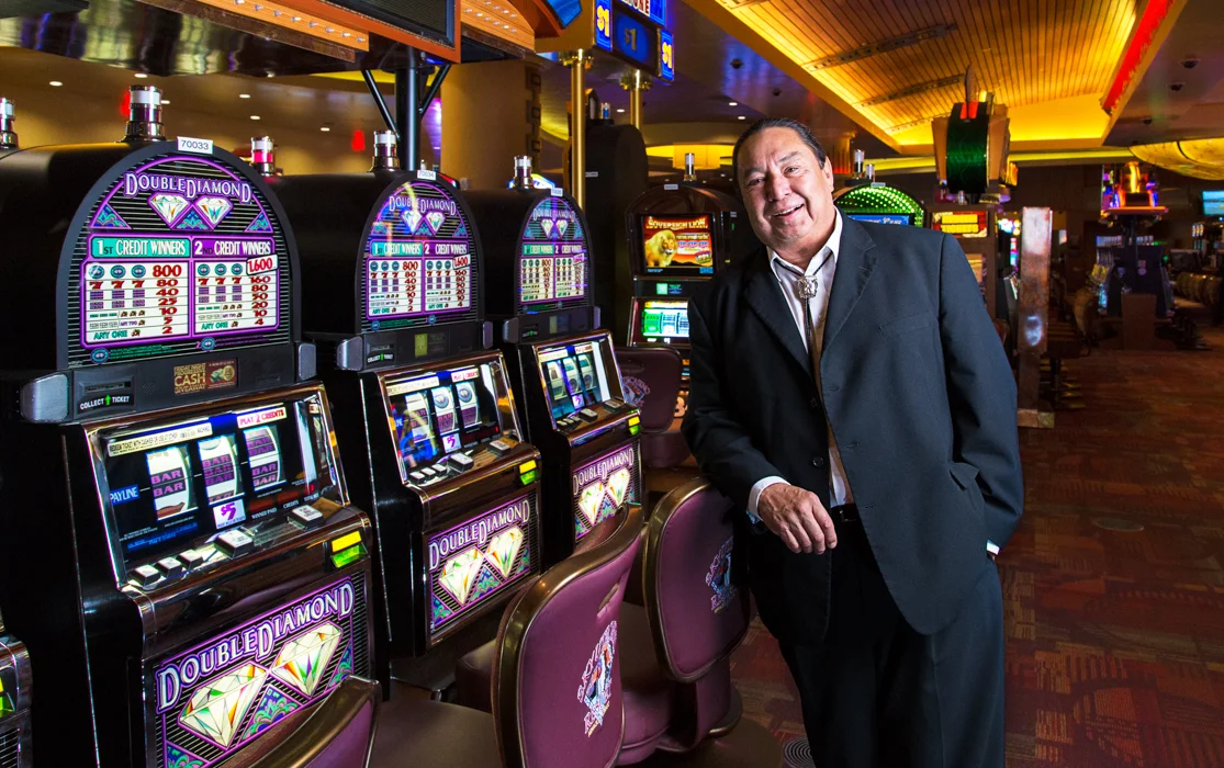 A man in a black suit standing next to slot machines in a casino, smiling.