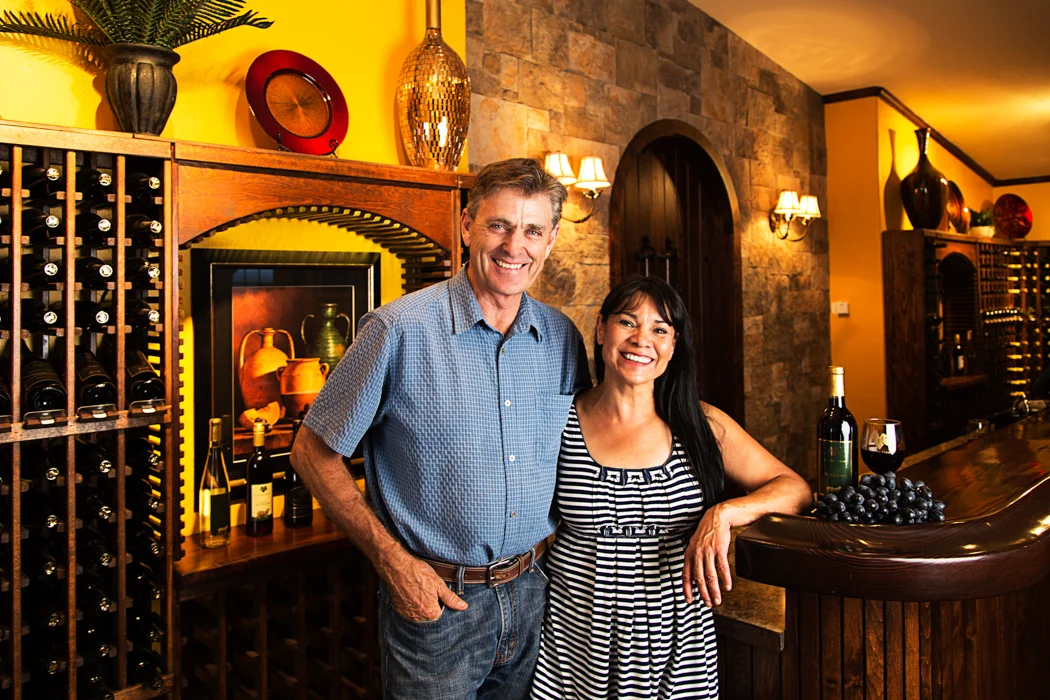 A smiling middle-aged man and woman standing behind a wine bar with bottles of wine, grapes, and a wine bottle on the counter in a warmly lit wine cellar or tasting room.