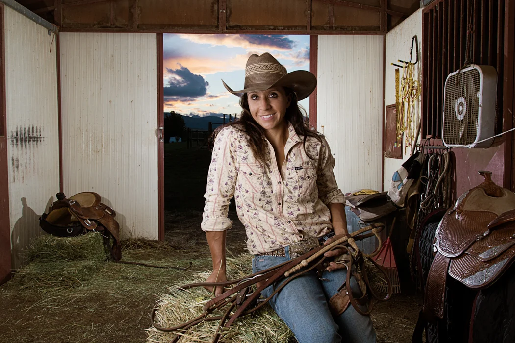 A woman in cowboy attire and a wide-brimmed hat sitting in a barn with western saddles and tack, holding a riding crop, during sunset.