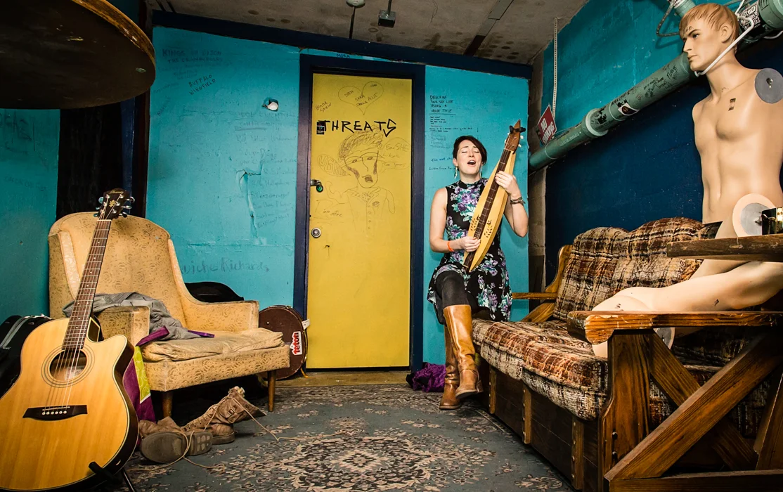 A woman singing and playing a musical instrument in a cluttered room with guitars, a mannequin, and colorful walls.