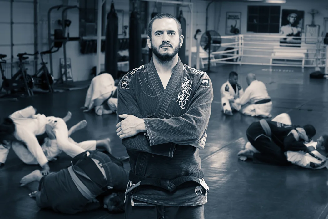 A man in a Brazilian Jiu-Jitsu gi stands with arms crossed in the foreground of a martial arts gym, with students practicing techniques on the mats in the background.