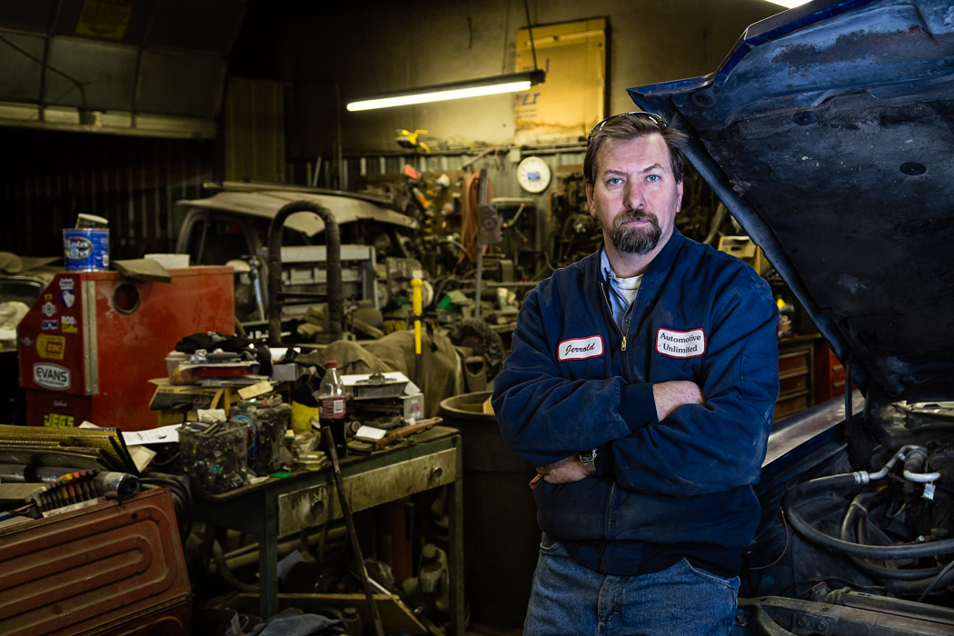 A man with a beard and mustache in a blue jacket standing in a cluttered mechanic workshop with open hood of a vehicle on his right.