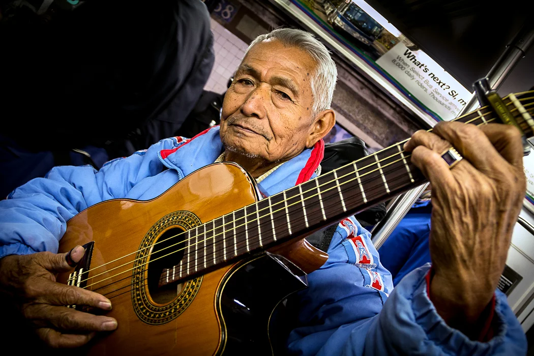 An elderly man with gray hair, wearing a blue jacket, is playing an acoustic guitar on a subway train. He is looking directly at the camera with a serious expression.