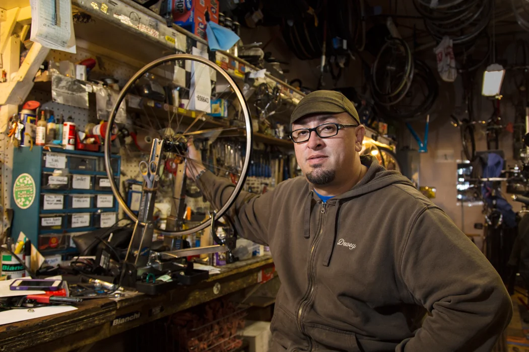 A man wearing glasses, a hat, and a brown hoodie with the name 'Dwayne' is working in a bicycle repair shop. He is holding a bicycle wheel with tools and bike parts on a cluttered workbench and shelves filled with bicycle parts and equipment in the b