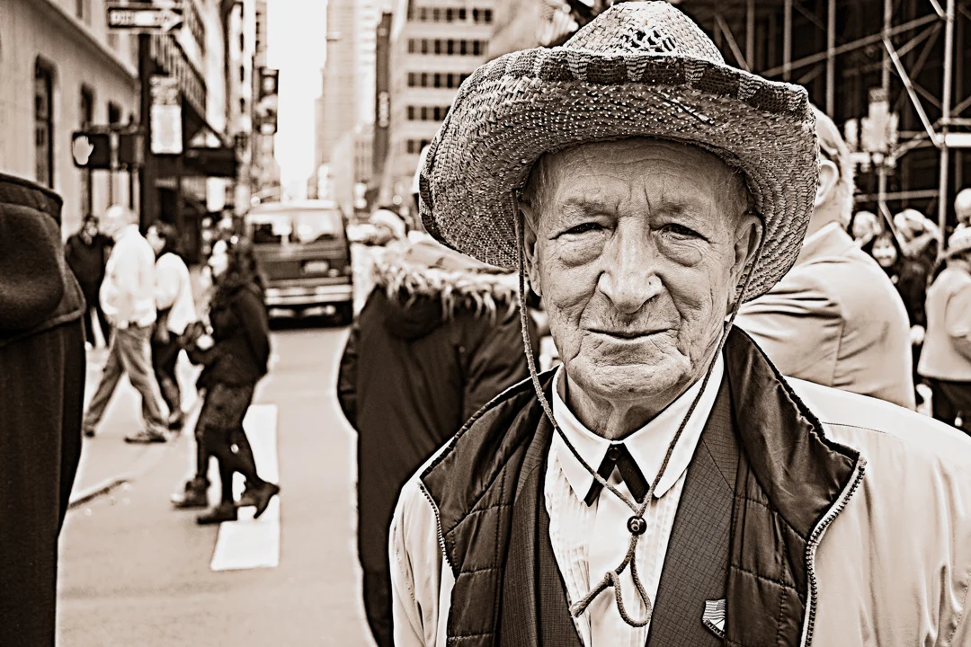 An elderly man wearing a wide-brimmed hat, standing on a busy city street among pedestrians and buildings.