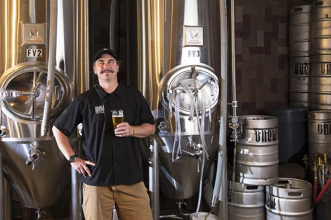 A man with a mustache holding a glass of beer, standing in front of stainless steel brewing kettles and tanks in a brewery.