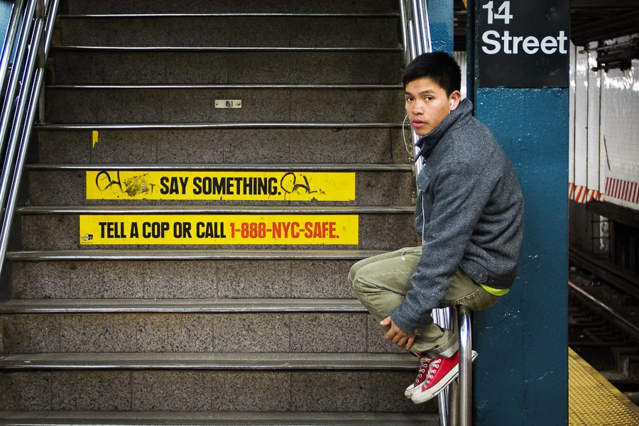 A young boy sits on a subway stair rail next to a sign for 14th Street. The stairs have yellow safety warnings and a sign with urging to say something or call a safety hotline.