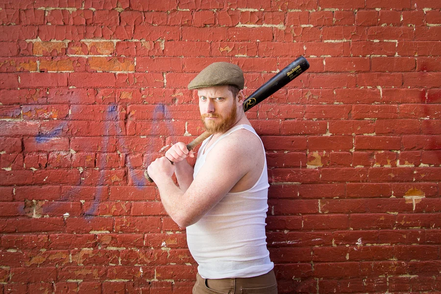 A man with a beard and red hair wearing a flat cap and a white sleeveless shirt, holding a baseball bat over his shoulder, standing against a red brick wall.