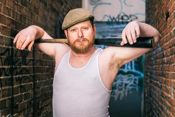 A man with a beard wearing a flat cap and a sleeveless shirt, holding a baseball bat over his shoulders in an alley with graffiti-covered walls.