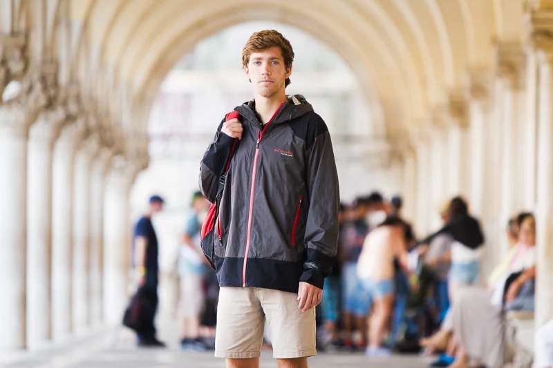 A young man with brown hair standing in a historical architectural passageway, wearing a dark gray and red jacket, beige shorts, and carrying a backpack.