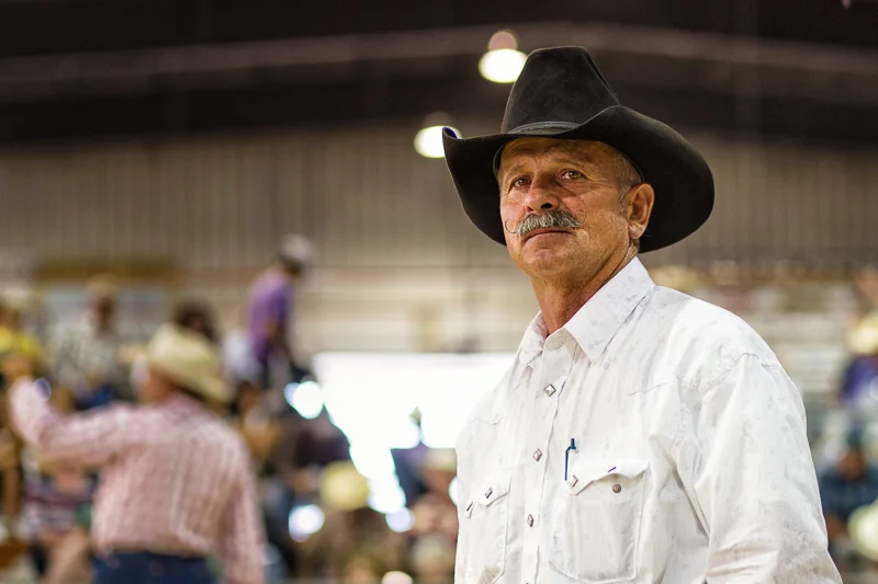 Older man wearing a cowboy hat and a white button-up shirt in a busy indoor setting with people in the background.