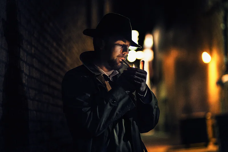 A man with a beard, wearing a hat and a dark jacket, lighting a cigarette in a dimly lit alley at night.