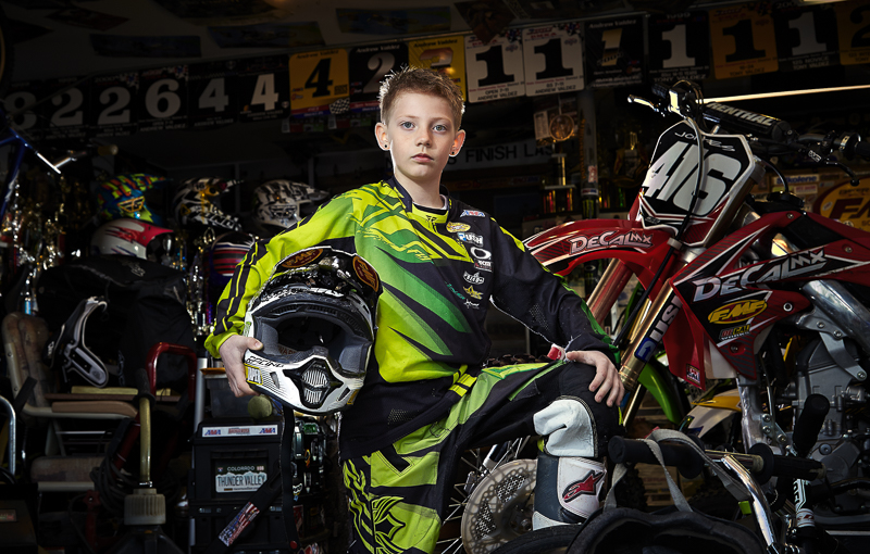 Young boy in racing gear sitting on a motocross bike in a garage with helmets and racing banners.