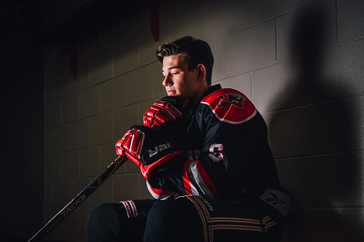Young male hockey player sitting against a wall, wearing a black and red hockey jersey and red gloves, holding a hockey stick, with a contemplative expression in a dimly lit setting.