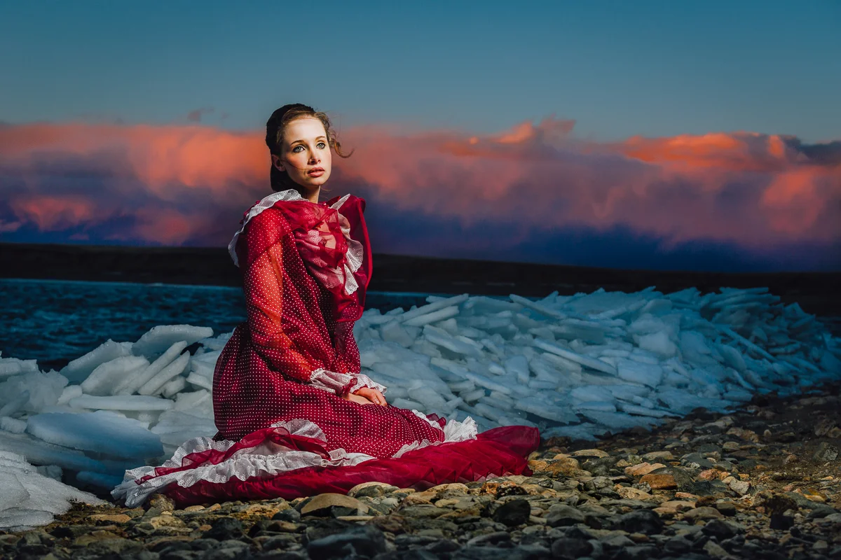 A woman in a red and white vintage dress sitting on a rocky shore by ice and water during sunset or sunrise.