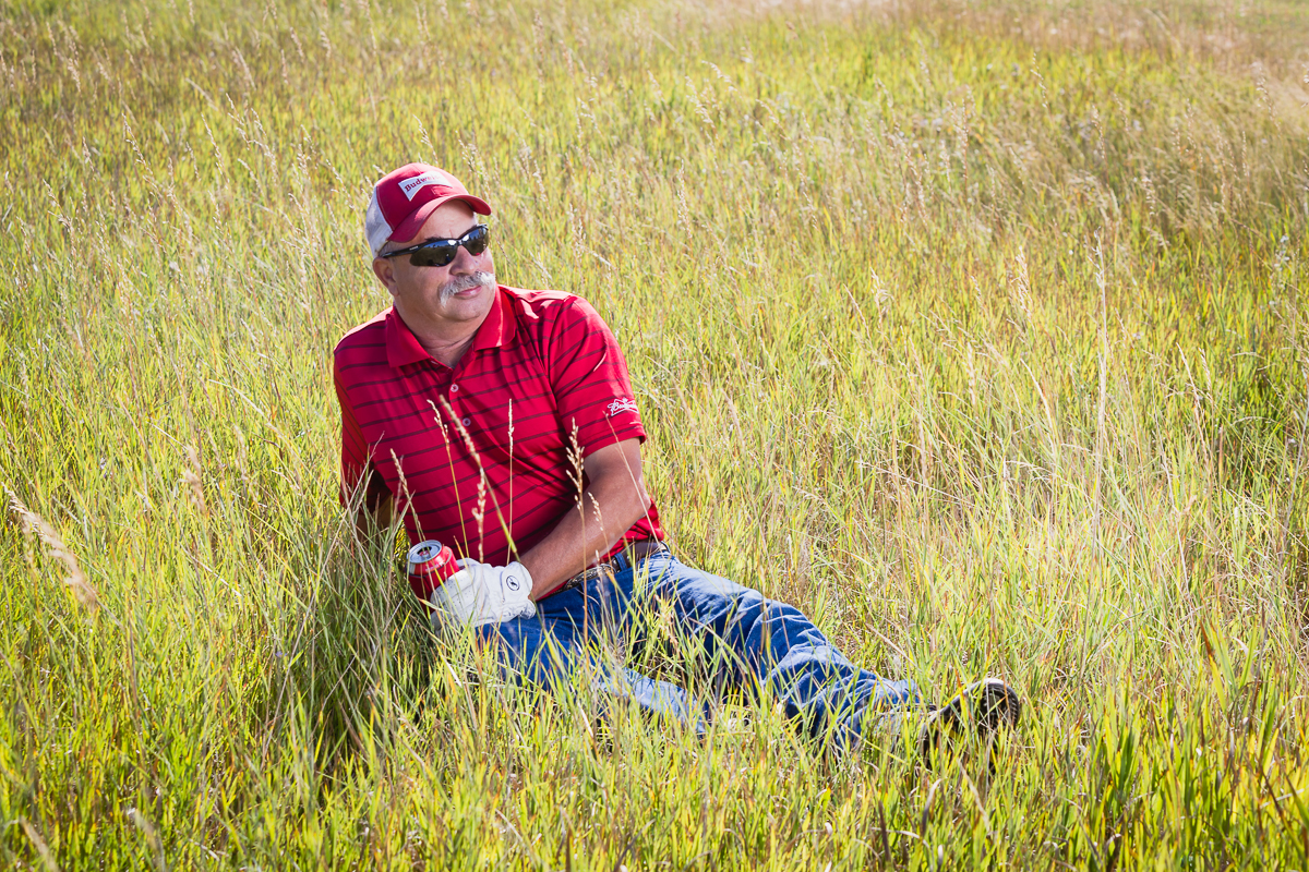 A man sitting in tall grass in a field, wearing sunglasses, a red cap, a red striped polo shirt, blue jeans, and holding a can of soda, relaxing on a sunny day.
