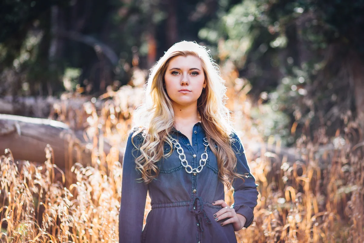 A young woman with long, wavy blonde hair standing outdoors in a forested area during daytime.